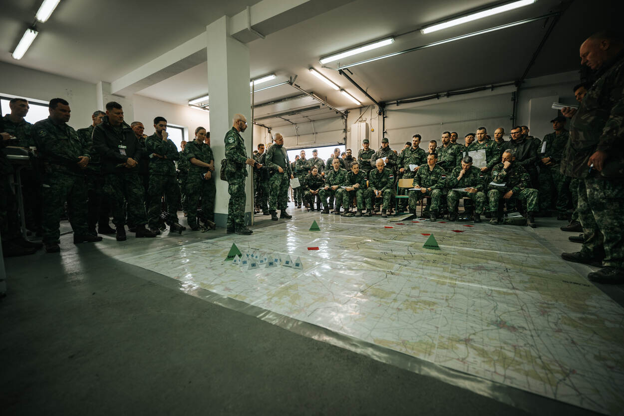 Alle aan de oefening deelnemende militairen staan in een loods rond een grote kaart, die op de grond ligt. Twee militairen geven uitleg over wat er de komende dagen gaat gebeuren op welke locaties.