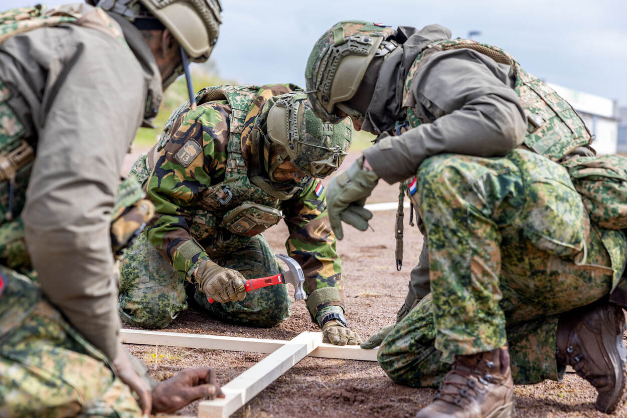 Drie militairen zitten rond een houten constructie. Een militair timmert met een rode hamer een spijker in de latten.