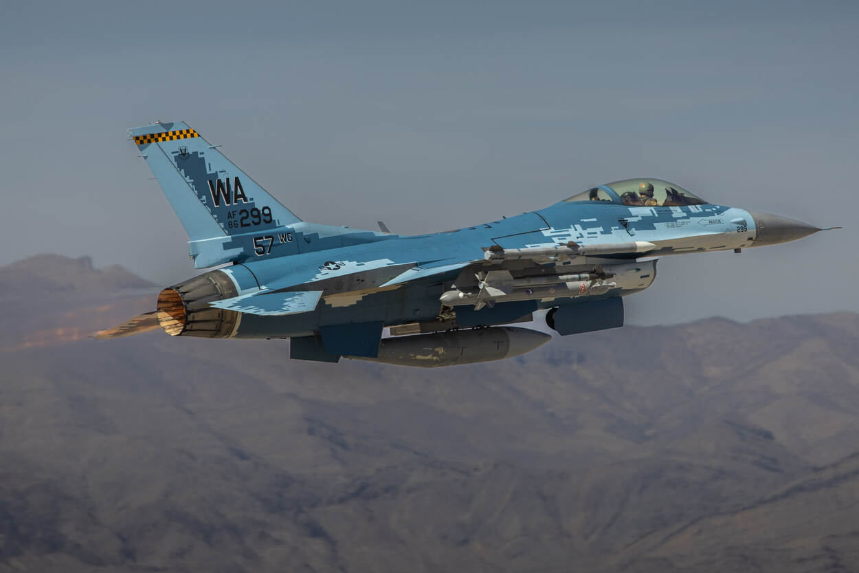 Een blauw/witte F-16 vliegend met onder meer een stoorpod onder de vleugel.