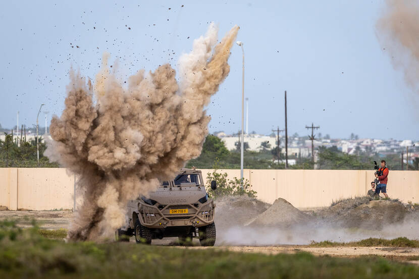 Een zandkleurig militair voertuig wordt geraakt door een explosie waarnaar grote stofwolken opstijgen.