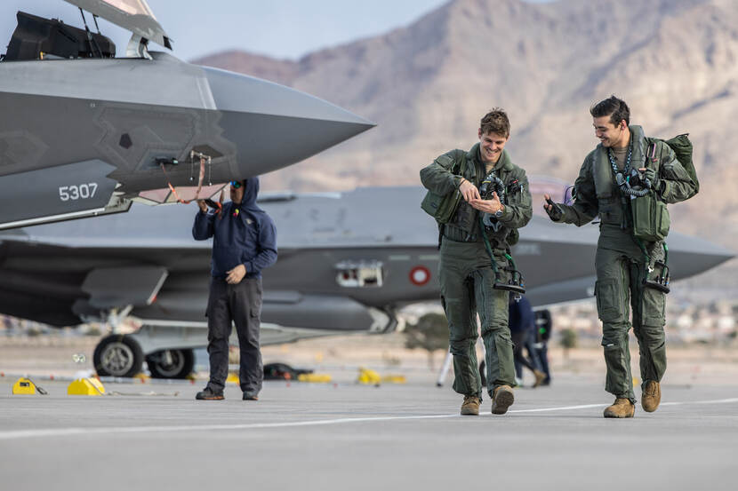 twee vliegers na afloop van een vlucht die naast elkaar lachend en gebaren makend naast elkaar lopen. Op de achtergrond staan enkele F-35’s.