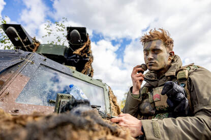 Een militair brengt camouflage aan naast zijn Fennek.