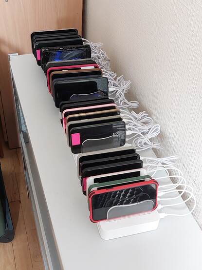 Ruim 20 telefoons in verschillende condities liggen naast elkaar en zijn ieder gekoppeld aan een kabel.