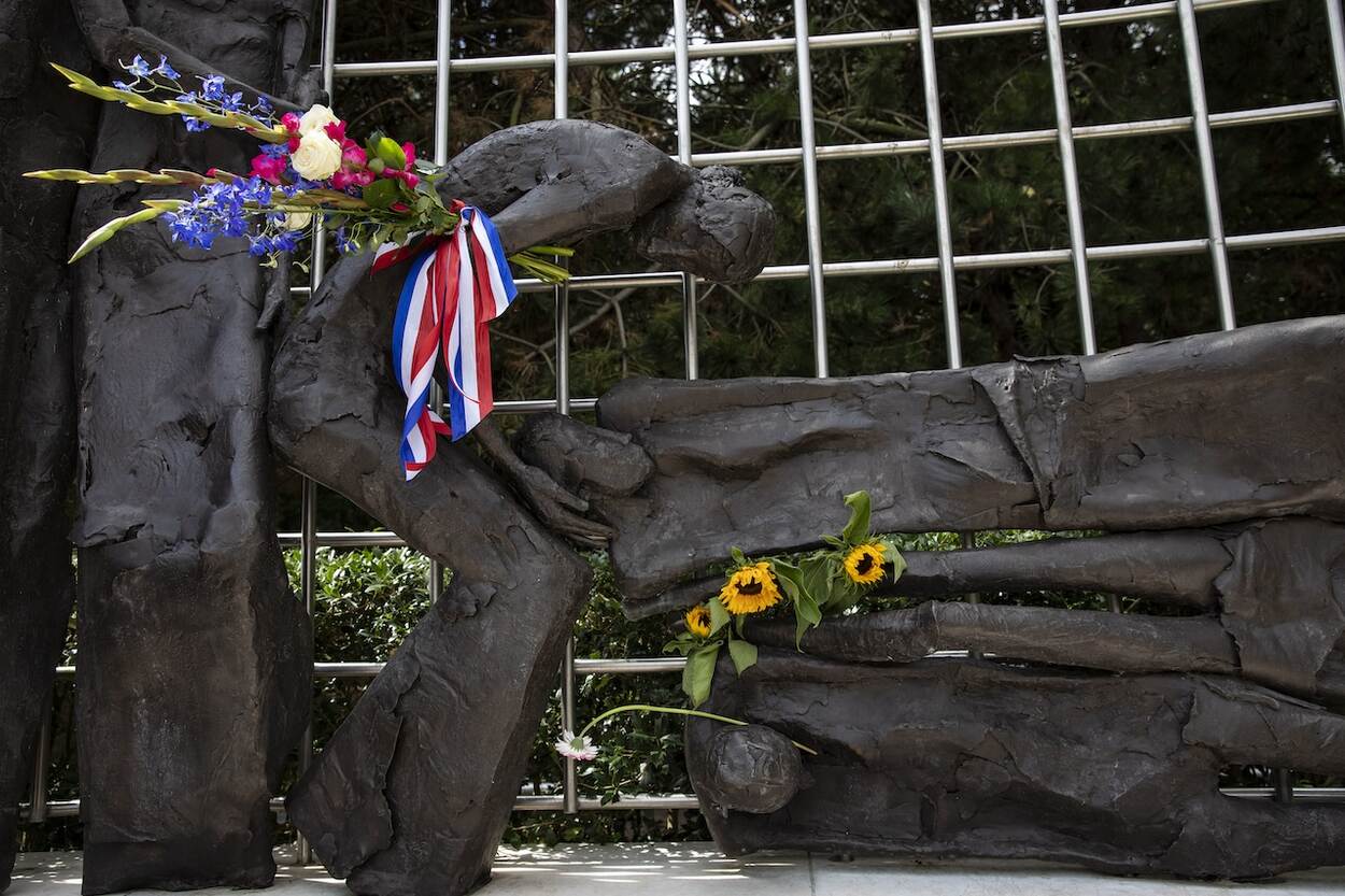 Foto van het Indisch Monument in brons, van een vrouw die voorovergebogen het hoofd vasthoudt van iemand die ineengekrompen op de grond ligt. Monument versierd met bloemen.