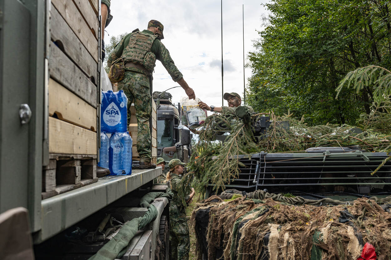 Een militair op een vrachtwagen deelt water uit aan de bestuurder van een gecamoufleerde Fennek verkenningsvoertuig in de zogenoemde bevo-straat.