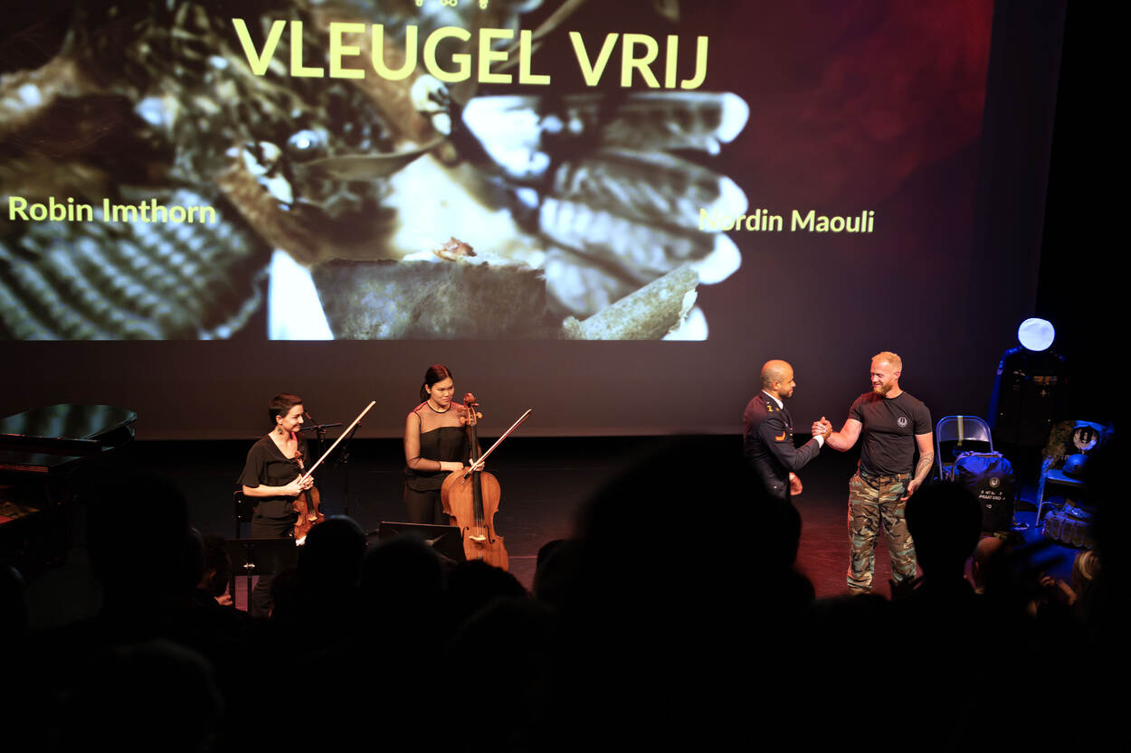 Twee mariniers schudden elkaar de hand op het podium van een theater, gezien vanachter de toeschouwers.