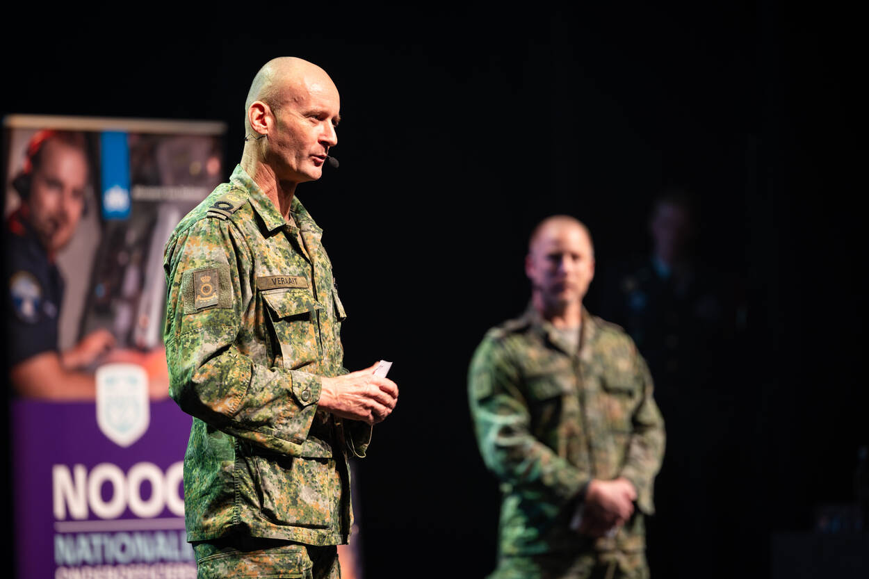 Krijgsmachtadjudant Mark Veraart staat in zijn gevechtspak op het podium in Nijkerk. Vaag achter hem staat een andere militair.