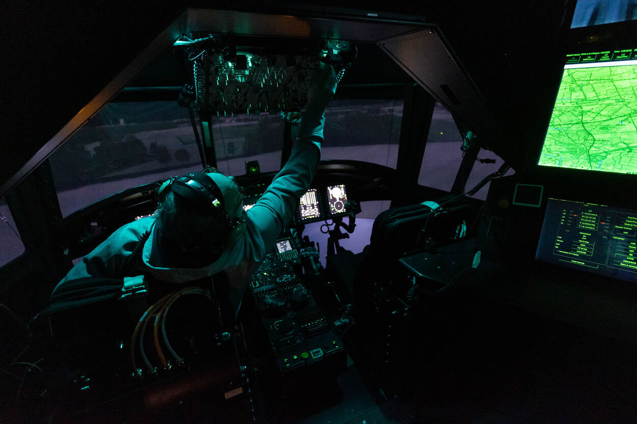 Een vlieger zit in een duistere vliegsimulator van een helikopter. De militair reikt met de rechterhand naar een knopje boven hem. In de hoek is een verlichte kaart van de omgeving te zien