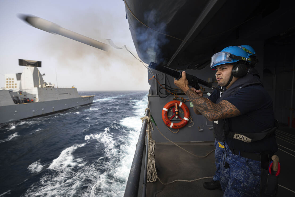 Een militair aan de Karel Doorman, aan de zijkant van het schip, schiet een brandstoflijn uit naar een schip, dat naast hem vaart.