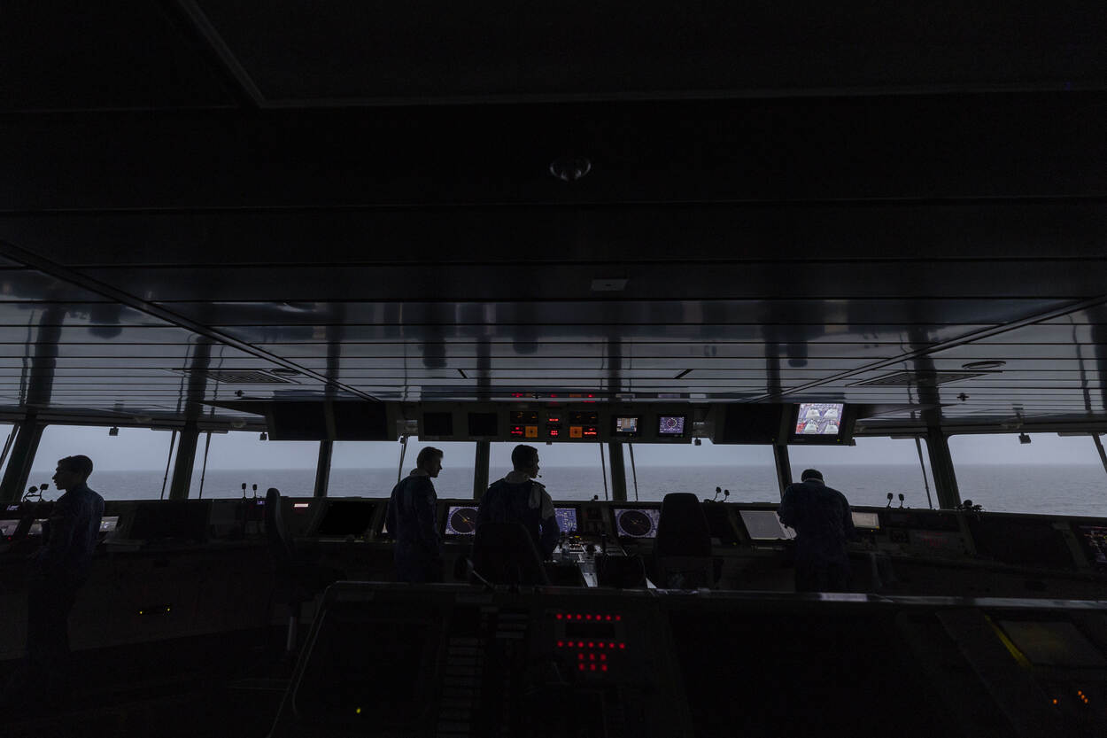 De brug aan boord van de Karel Doorman. Door de lichtval zijn enkel silhouetten van de marinemensen te zien. Op de achtergrond is de zee te zien.