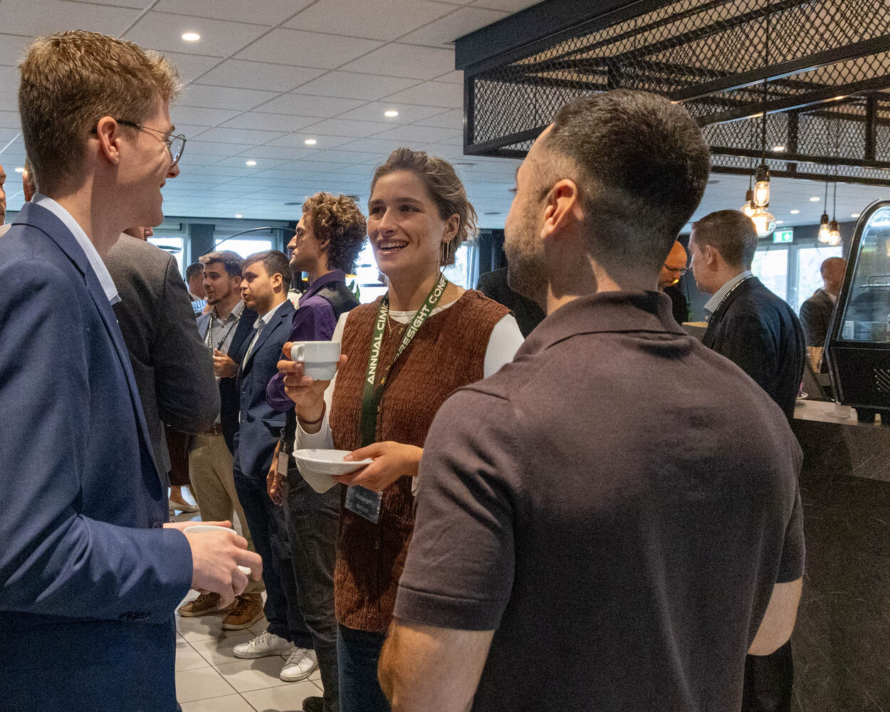 Drie mensen staan gezellig te kletsen onder het genot van een kop koffie met op de achtergrond meerdere mensen.