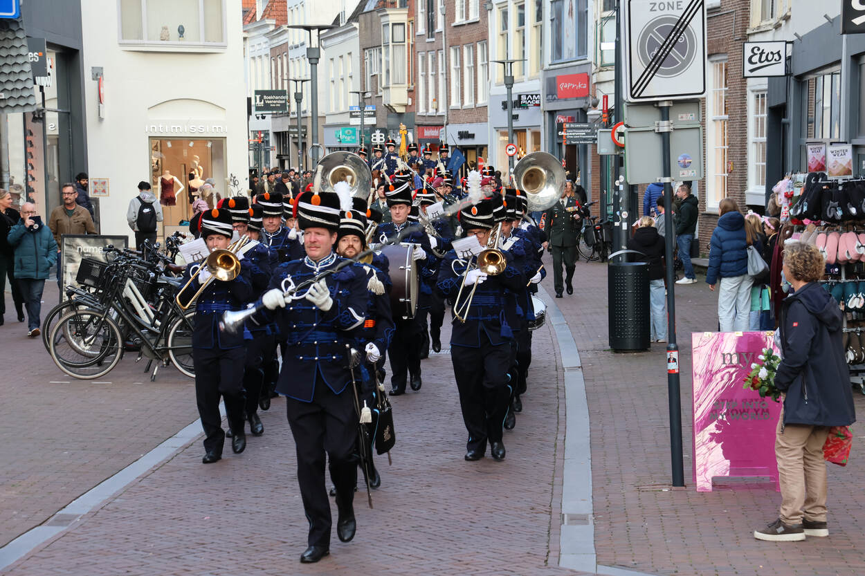 Het fanfareorkest in uniform en hoeden, spelend op koperblaas- en slagwerkinstrumenten, terwijl mensen langs winkels toekijken op een druk winkelgebied. Rechts: Een groep mensen in militaire kleding staat in gelid, met historische gebouwen en een kerktoren op de achtergrond onder bewolkte lucht.