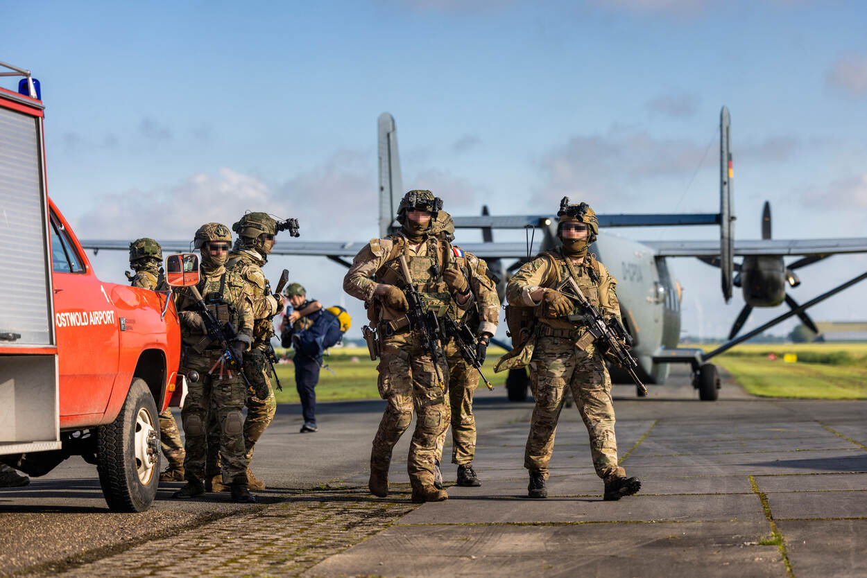 Bewapende miltairen lopen langs een landingsbaan, met achter hen een vliegtuig.