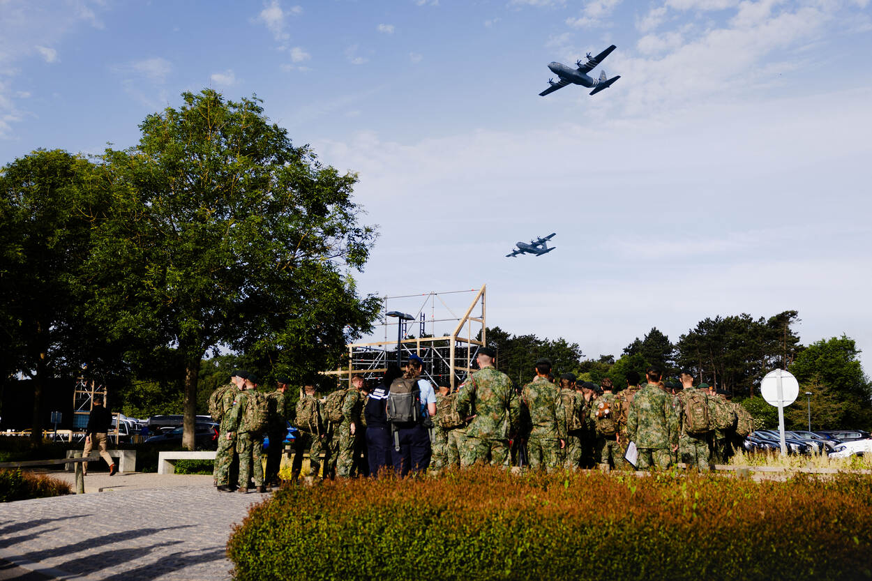 Een groep militairen staat voor de ingang van de Amerikaanse begraafplaats terwijl twee Amerikaanse, militaire vliegtuigen overkomen.
