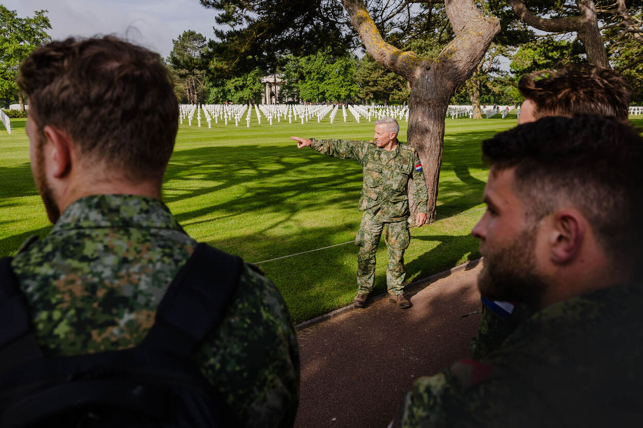 Een sergeant-majoor wijst naar de vele witte kruizen op de begraafplaats, terwijl collega’s naar hem luisteren.