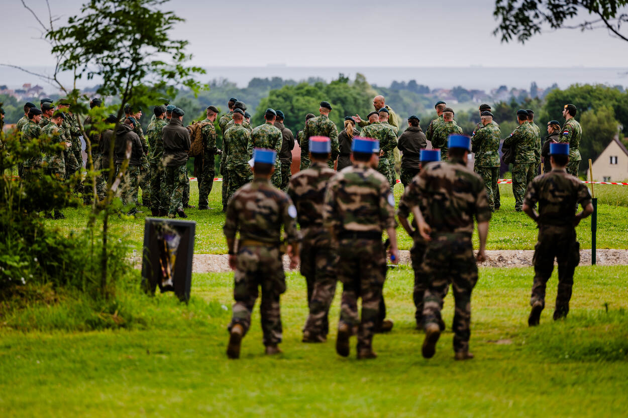 Op de achtergrond een groep Nederlandse militairen die in een kring naar een presentatie luistert, daarvoor een groep militairen met ronde, blauw-rode hoofddeksels.