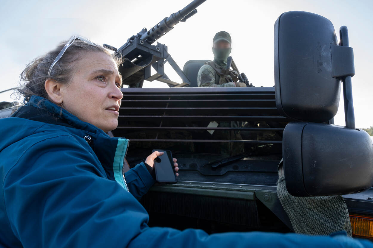 Een dame met een blauwe jas kijkt opzij, staand voor een Fennek-verkenningsvoertuig. Boven haar torent een militair met balaclava boven het voertuig uit.