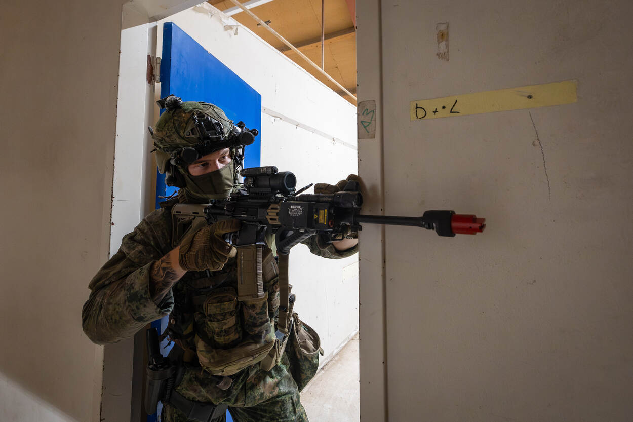 Militair met wapen staat in deuropening en kijkt gang in.