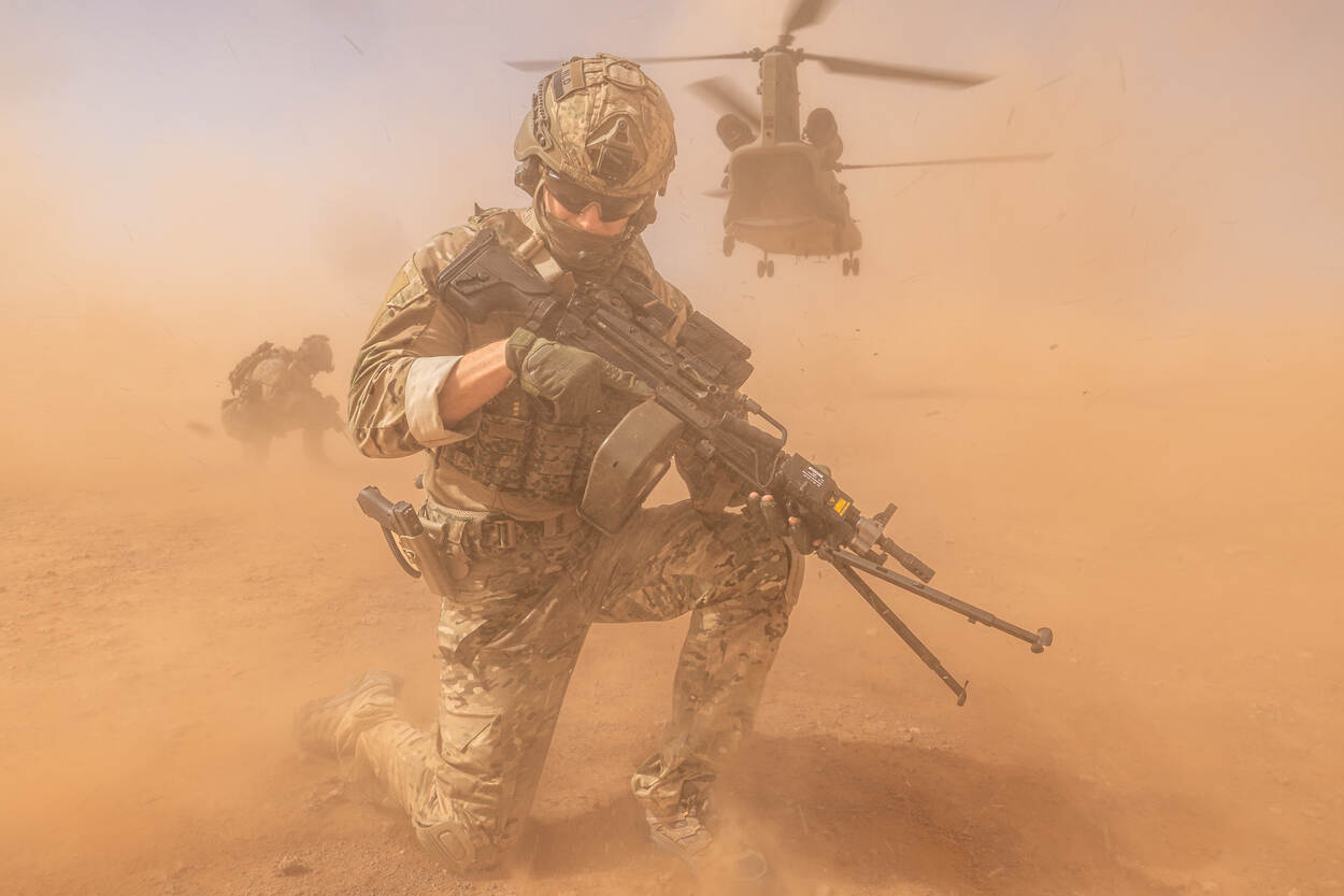 Een bewapende militair knielt, terwijl de landende heli voor opstuivend zand zorgt.