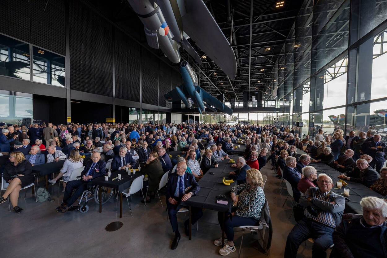 Grote hal met teintallen bezoekers zittend aan lange tafels. Oude vliegentuigen hangen aan het plafond.