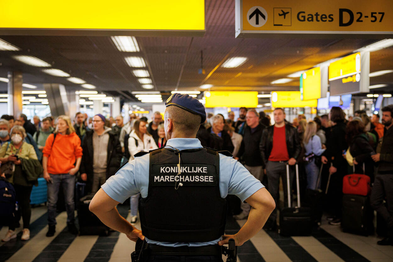 Een marechaussee, op de rug gezien, houdt op Schiphol een menigte reizigers in de gaten.