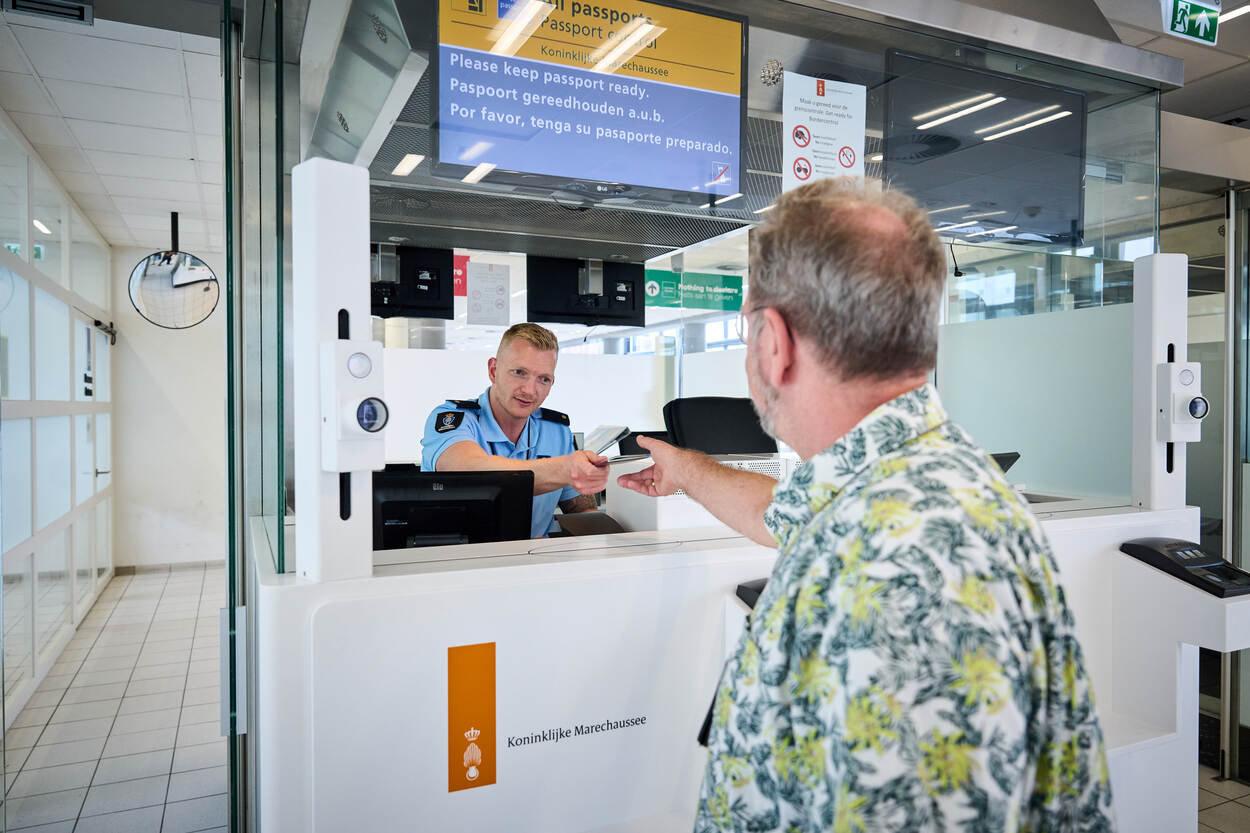 Een marechaussee geeft een reiziger zijn paspoort terug. Bij de balie hangt een speciale camera die wordt gebruikt voor het Entry/Exit System.