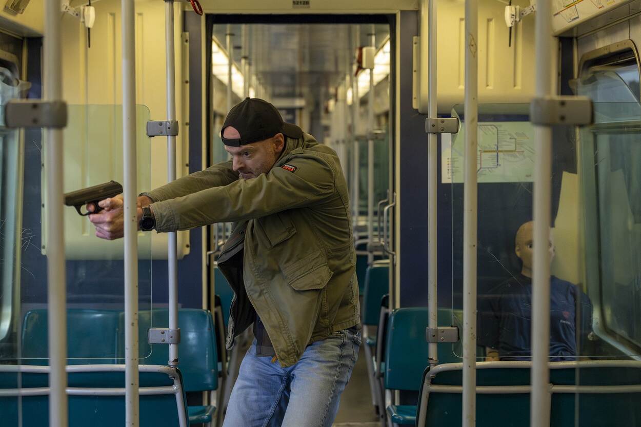 Man met petje op houdt met pistool in beide handen zienderogen iemand onder schot in de metro, op de achtergrond is een passagier zichtbaar.