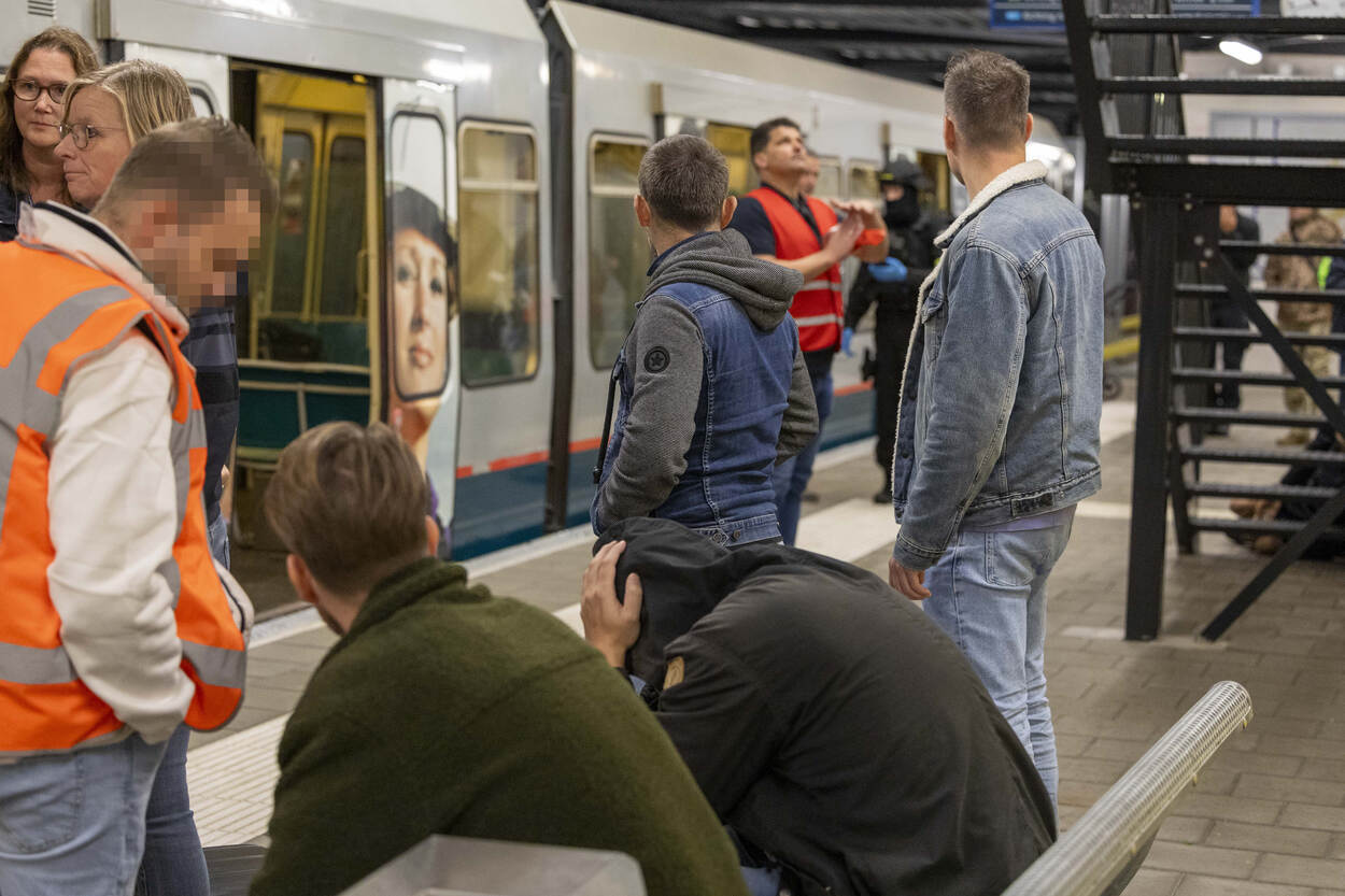 Twee mensen zitten op bankje metrostation, van wie een met capuchon op. Daaromheen een tiental mensen, van wie 2 met hesje en 1 in uniform KMar.