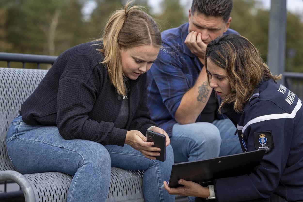 Man en vrouw op bankje laten iets op telefoon zien aan medewerker KMar met iPad.