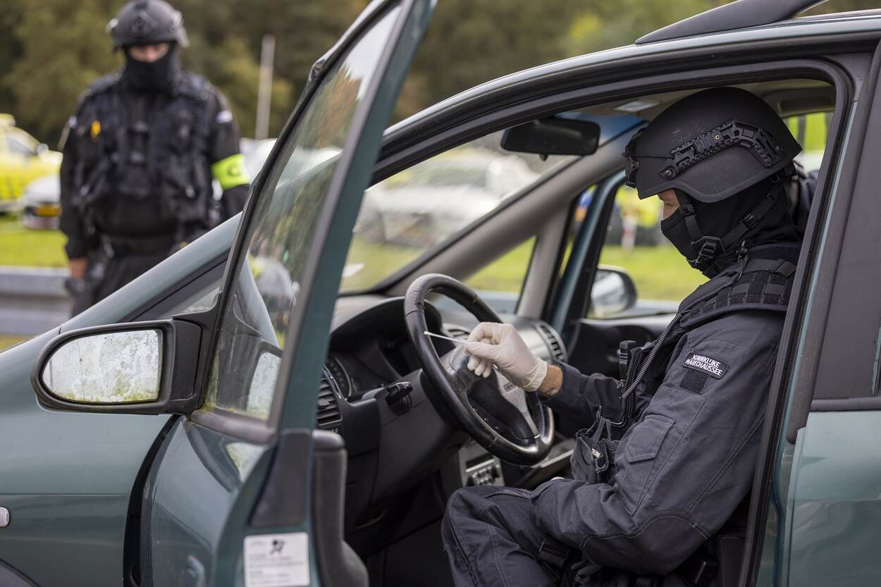 Medewerker KMar in tenue en helm op checkt auto met openstaande deuren op sporen. Op achtergrond een collega van KMar met helm op.