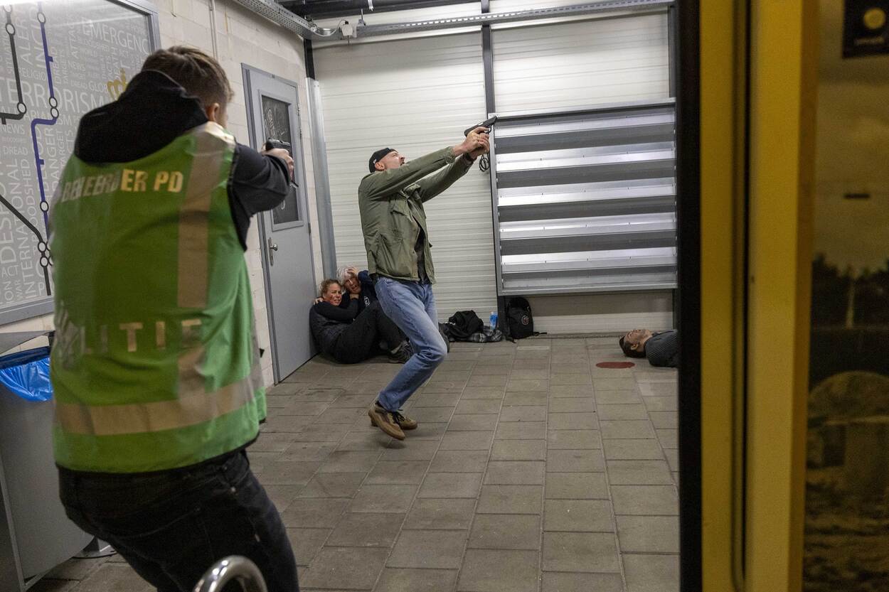 Politieman met hesje aan richt pistool op neerstortende man met pistool  en petje op. Links zitten 2 mensen terwijl rechts man ligt naast plas bloed.