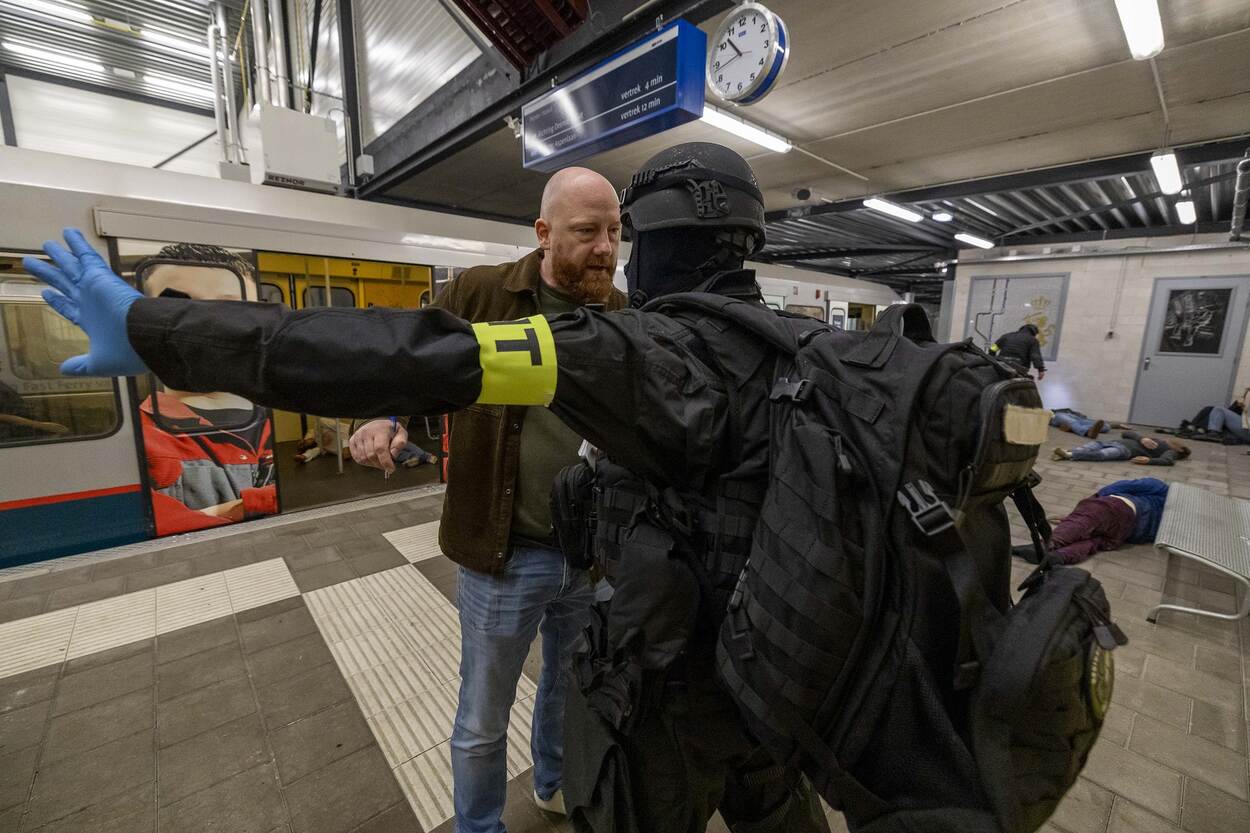 Lid van de KMar in tenue met helm in gesprek met kale man met baard. Op de achtergrond een metro met openstaande deuren en liggende mensen.