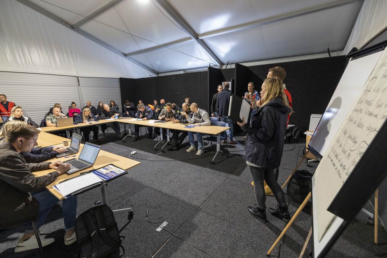 Mensen in tent in een u-vorm aan tafels met verschillende laptops voor zich. 2 personen staan en praten voor flipover.