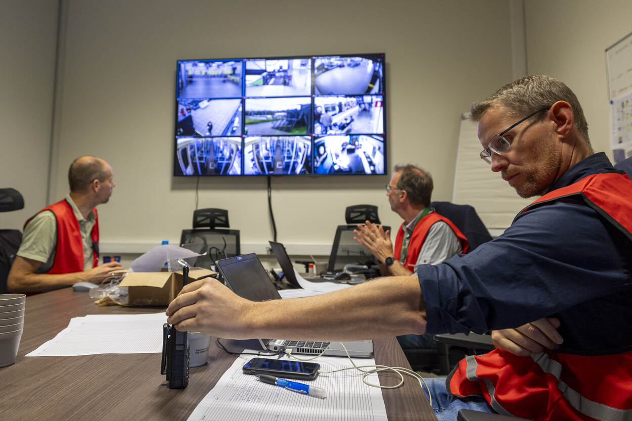 3 personen met hesje zitten in vergaderzaal aan tafel met laptops en telefoons met op de muur scherm met bewakingsbeelden.