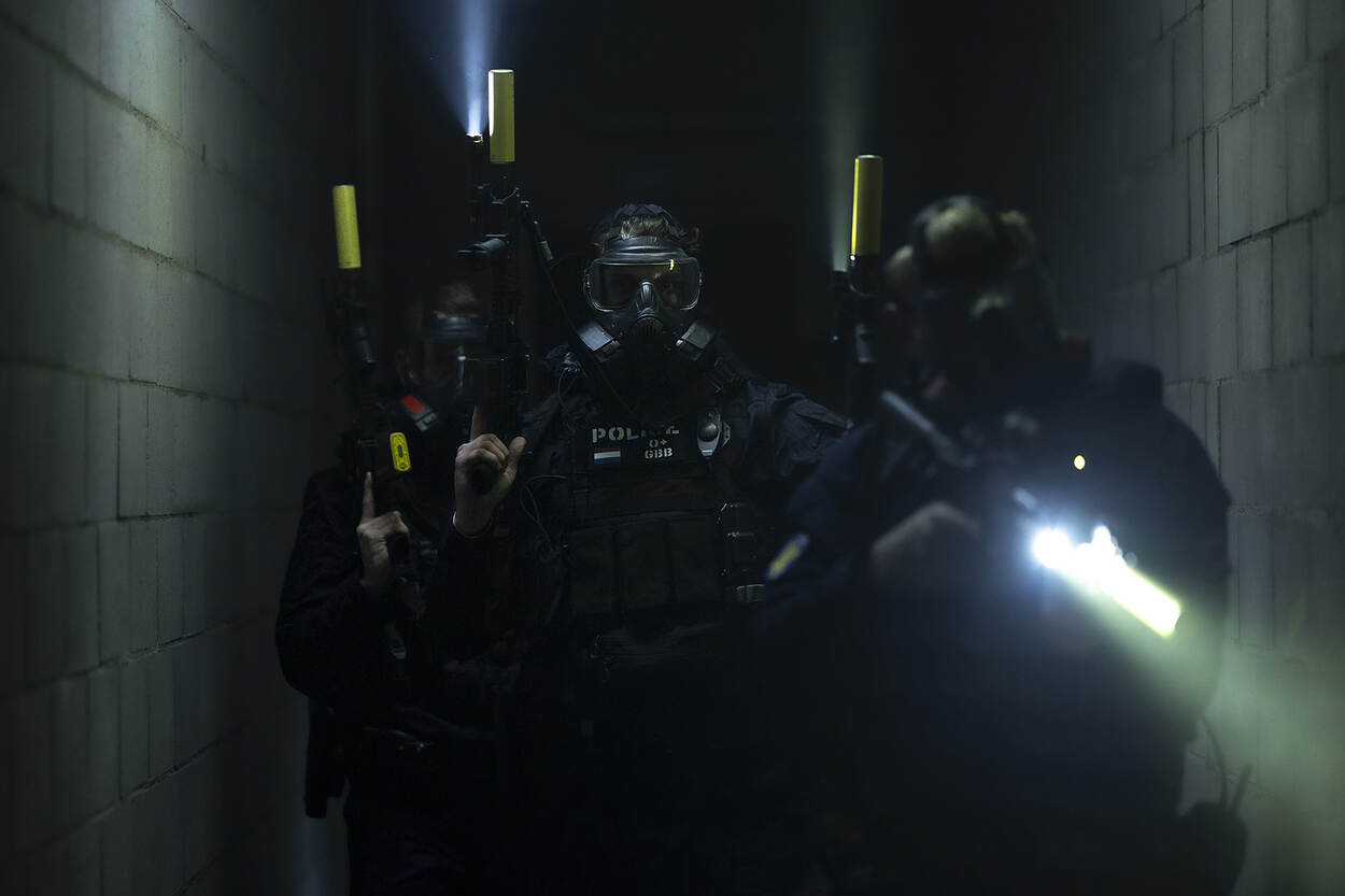 Drie gemaskerde GBB’ers lopen met getrokken wapen door een gang.