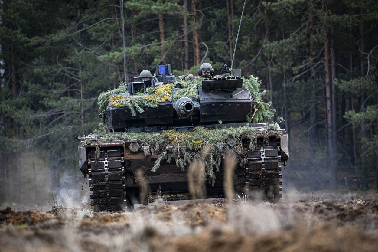 Met dennentakken gecamoufleerde Leopard 2A6 op zanderig terrein met twee militairen uit de koepel.