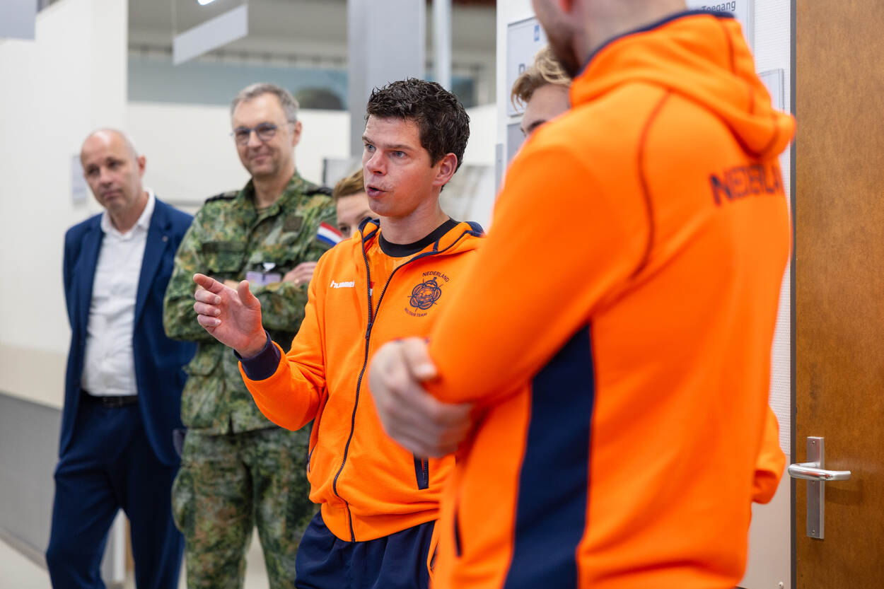 Vier mannen in een binnenruimte: twee in feloranje jassen met "Nederland" erop, een in militair uniform, en een in pak; de voorste man praat met gebaren.