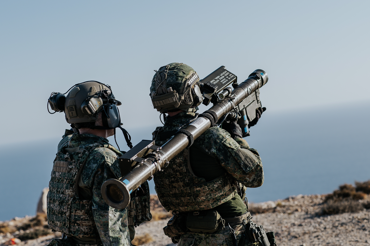 een schutter richt zijn lanceerbuis op een doel. Een instructeur kijkt mee.