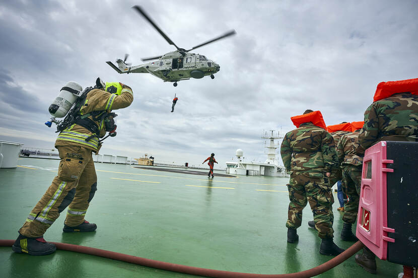 Een militair daalt vanuit de helikopter af aan boord van een schip. In beeld ook een groepje militairen en een brandweerman.