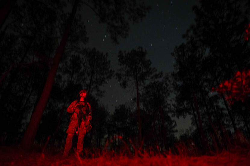 Een hele donkere foto met een zichtbare sterrenhemel waarop een militair met een rood lampje is uitgelicht.