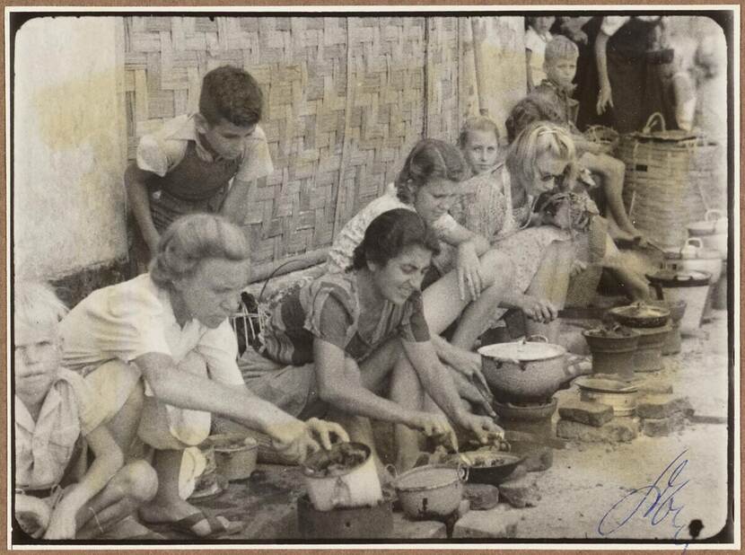 Zwart-wit foto van een rij kokende vrouwen. Links achter de vrouwen staat een jongetje mee te kijken.