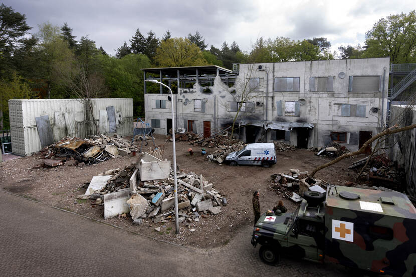 Voorzijde van het nieuwe oefengebouw in Hilversum voor militaire genezerikken. De muren zijn weggeslagen, te midden van de brokstukken staat een gesloopte ambulance.