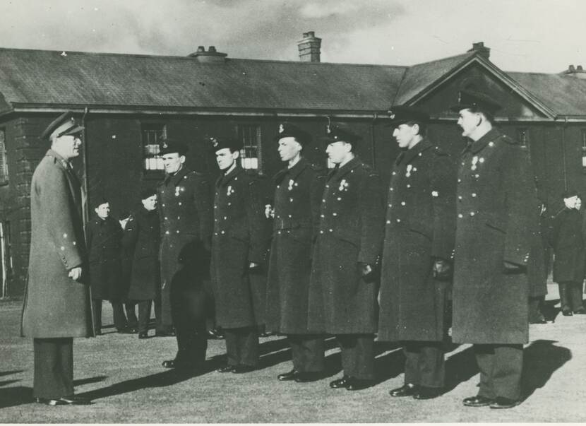 Zwart-witfoto van een rijtje militairen in lange zwarte jassen aan en zwarte petten op. Voor hen staat een officier met een grijze jas die hen een onderscheiding gaat uitreiken.