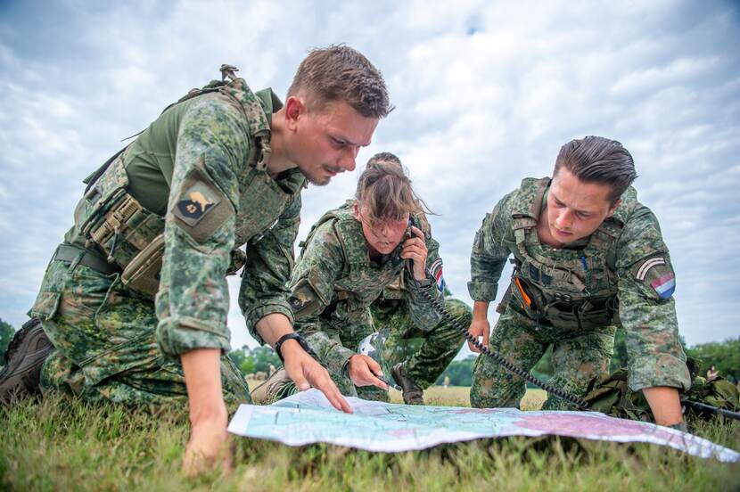 Drie militairen in uniform zitten geknield boven een kaart, waarbij een per telefoon gegevens doorstuurt.