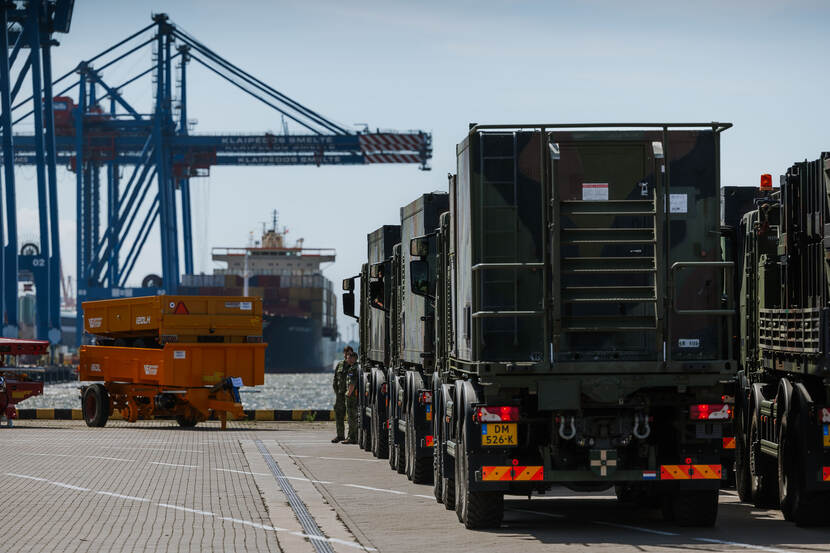 Militaire trucks staan op de kade, in de verte ligt een transportschip in de haven.