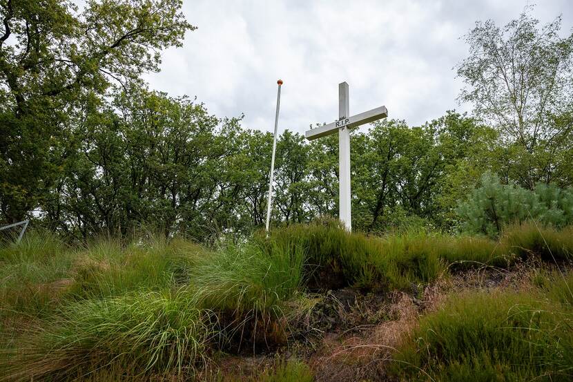 Een wit kruis met daarop de afkorting ‘R.I.P’, tussen het hoge gras. Daarnaast een lege vlaggenmast en op de achtergrond bomen.