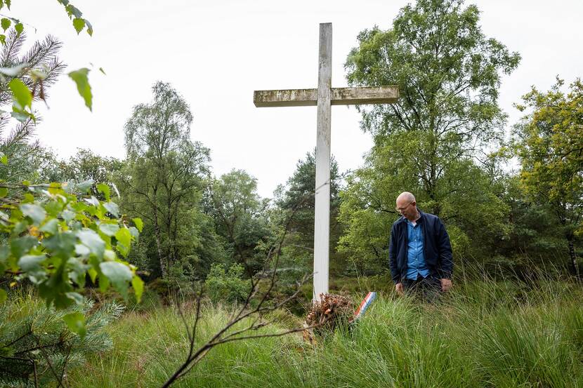 Van Loo naast het houten kruis. Hij kijkt naar een verdord boeketje van de herdenking op 4 mei.