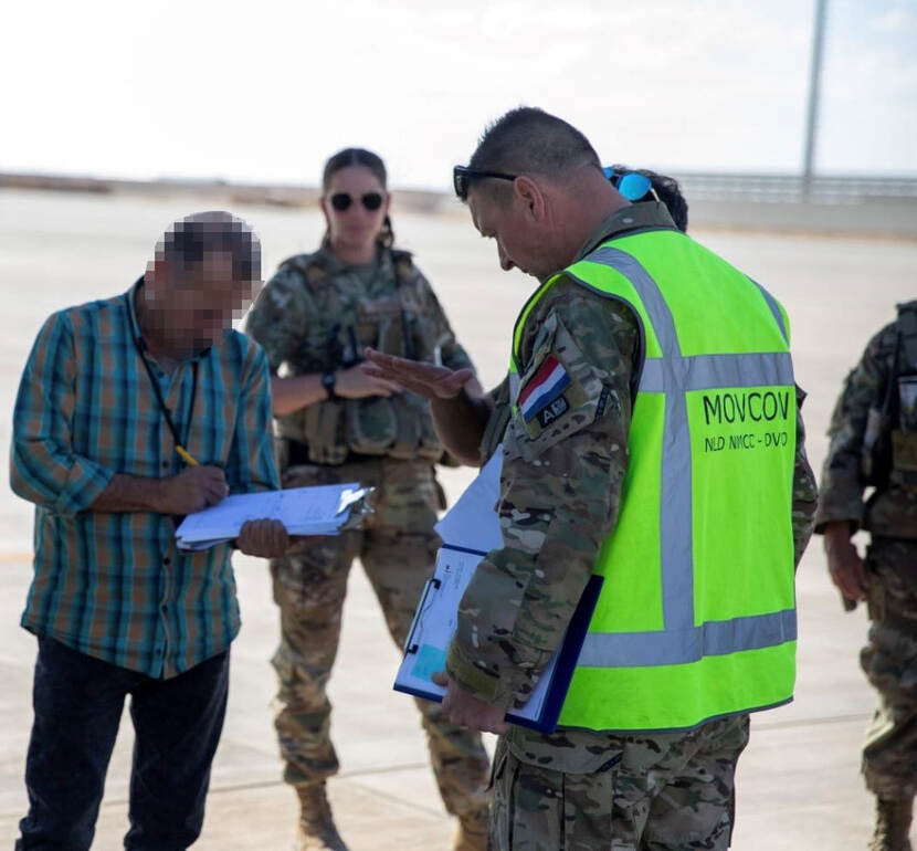 Een douanier in Irak buigt zich over de papieren in zijn hand, sergeant-majoor Dennis kijkt vanaf de overzijde mee. Op de achtergrond staan andere militairen.