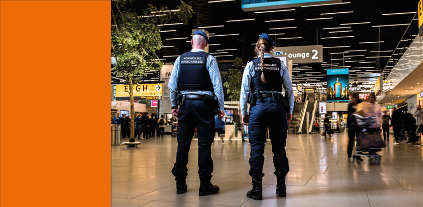 Twee collega’s van de marechaussee achter de douane op Schiphol, op de rug gefotografeerd.