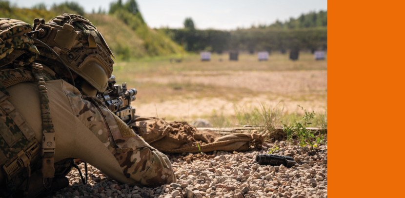 Militair ligt plat op z'n buik in het droge zand en kijkt door het richtmiddel van z'n lange wapen om in de verte het doel, een afbeelding op een kartonnen bord, te raken.